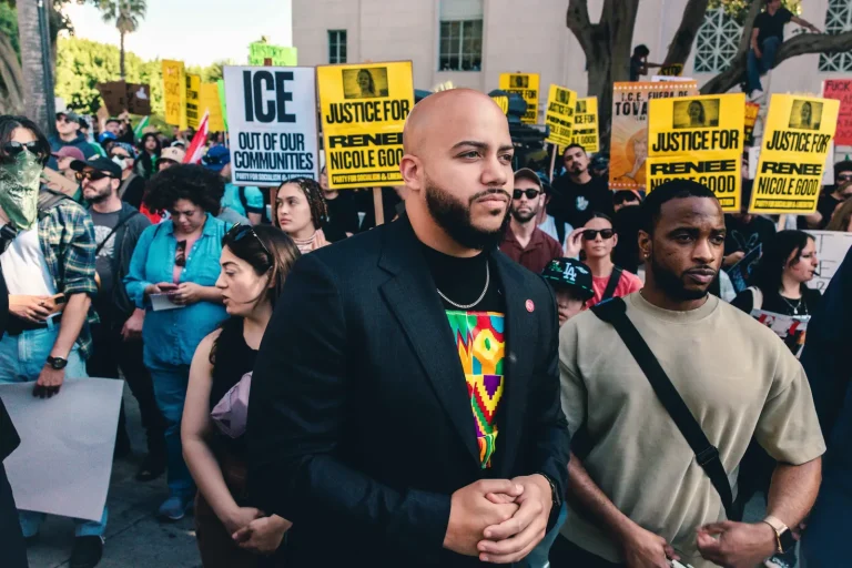 Isaac Bryan standing among demonstrators at an anti-ICE rally, hands clasped in front of him, with a sign reading ‘ICE out of our communities’ and rows of yellow placards raised behind him.