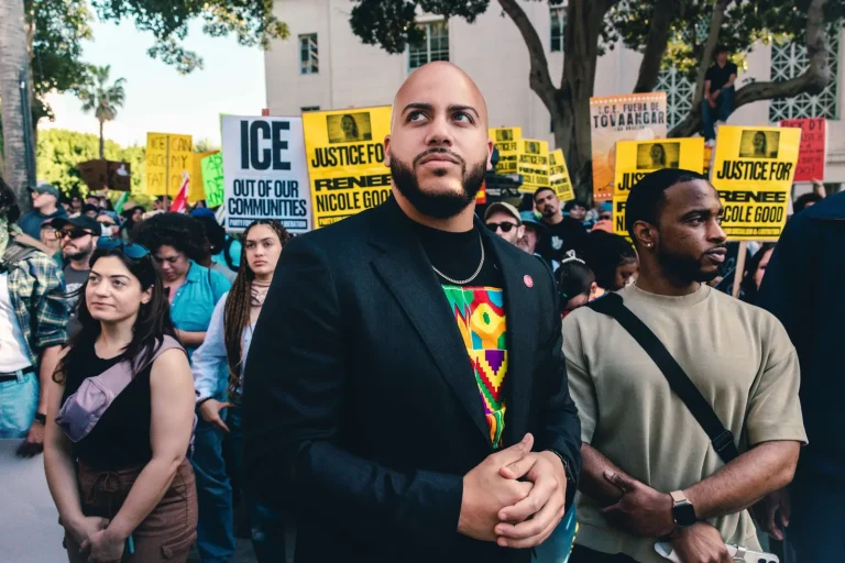 Isaac Bryan looking out over the crowd at an anti-ICE rally in a dark blazer over a kente-print shirt, an ‘ICE out of our communities’ sign and yellow placards behind him.