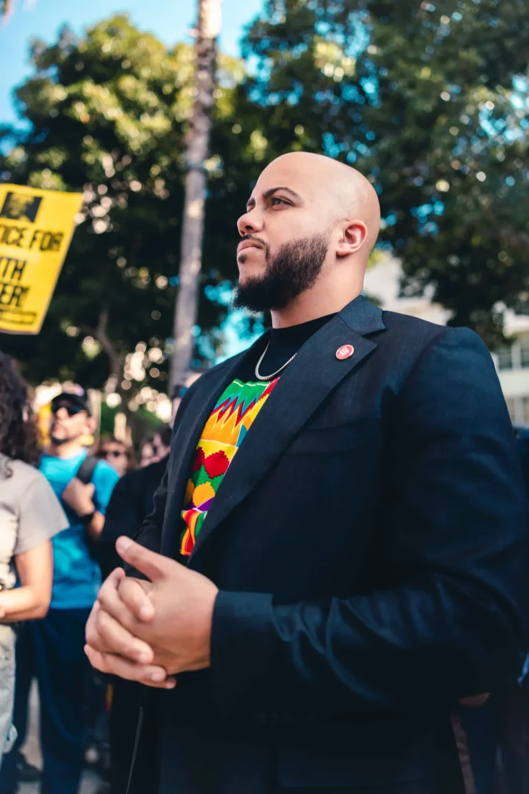 Isaac Bryan in profile at an outdoor rally, hands clasped in front of him, a palm tree and a protester’s yellow sign blurred in the background.