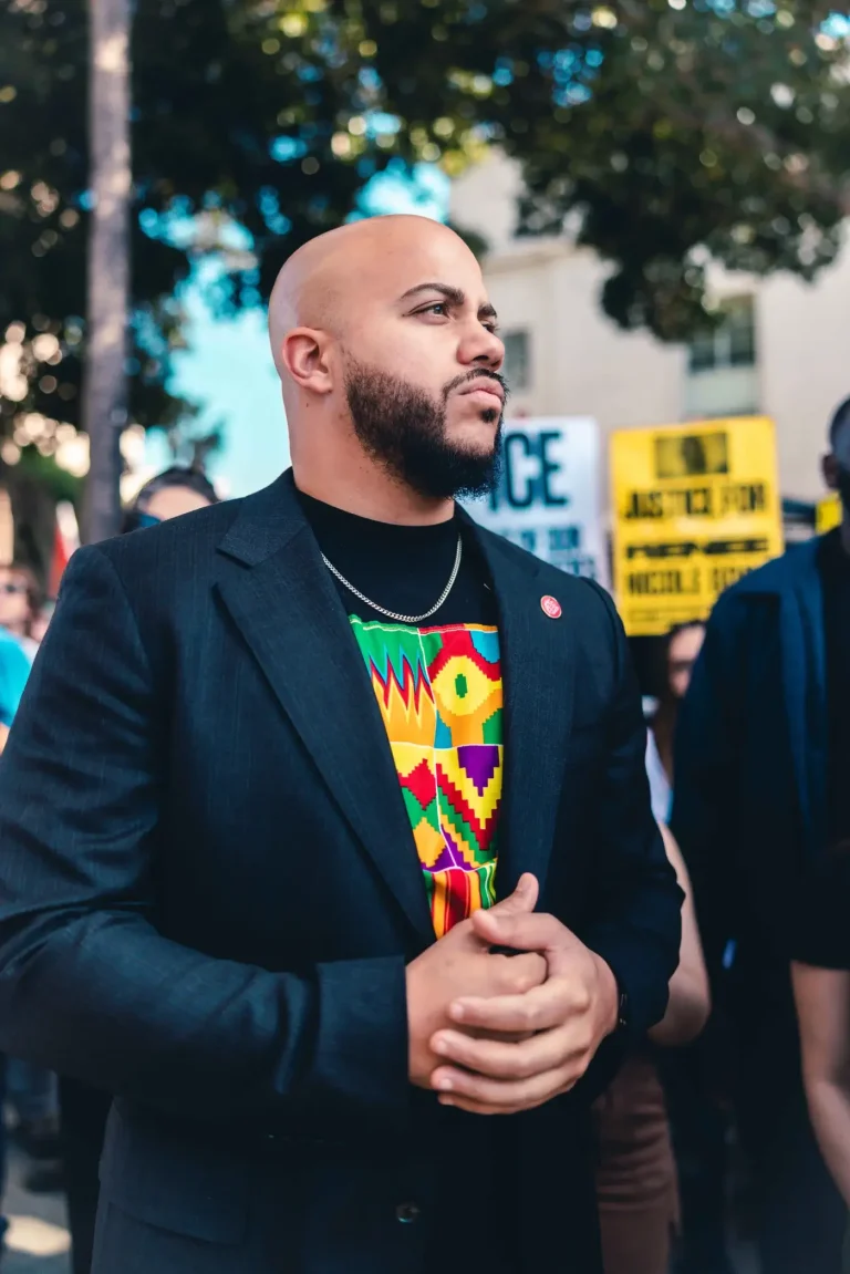 Isaac Bryan standing with his hands clasped at a rally, wearing a dark blazer over a brightly patterned kente-print shirt, protest signs out of focus behind him.