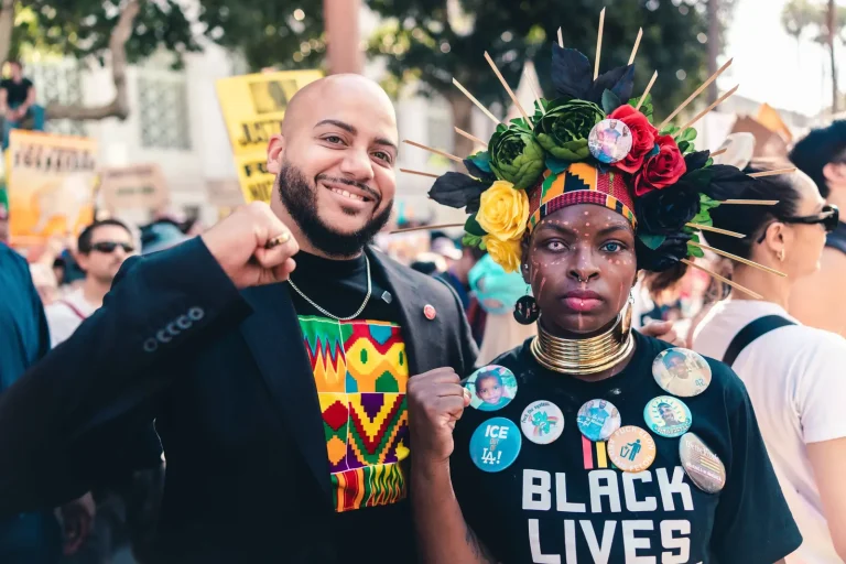 Isaac Bryan smiling with his fist raised beside a demonstrator wearing an elaborate floral and spiked headdress, a brass neck ring and a ‘Black Lives’ shirt covered in buttons.
