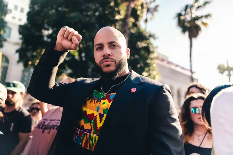 Isaac Bryan raising a clenched fist at an outdoor rally, palm trees and fellow demonstrators behind him in the afternoon sun.