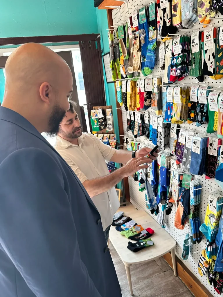 Isaac Bryan in a blue suit jacket visiting a small neighbourhood shop, looking over a pegboard wall of brightly patterned socks with the shopkeeper.