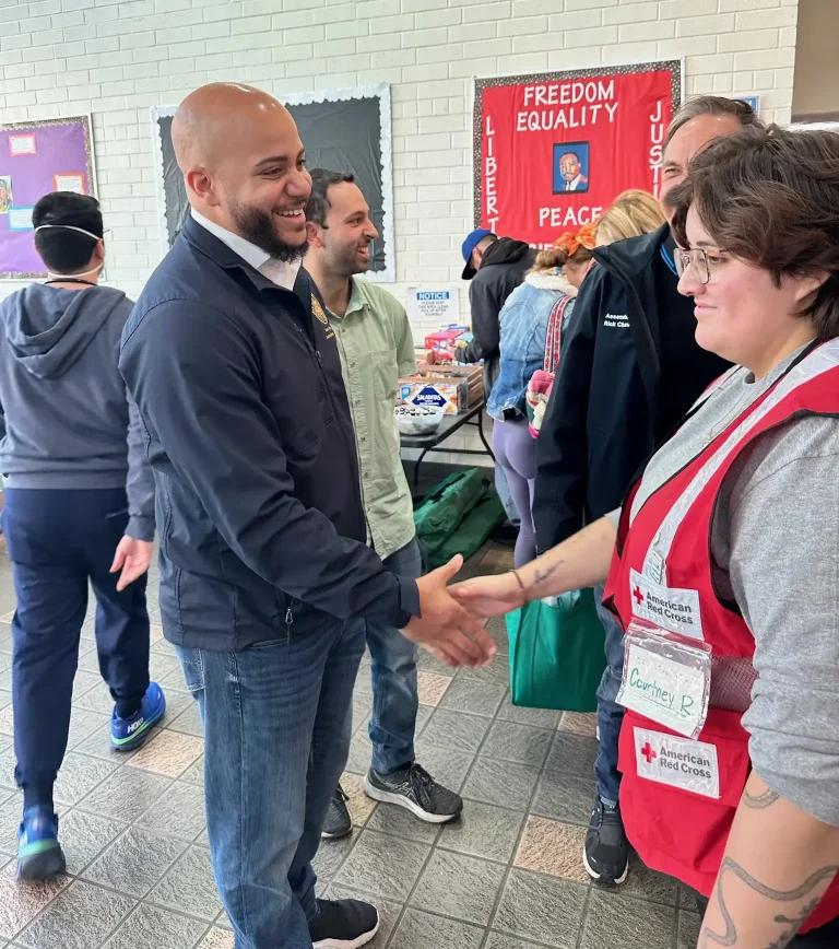 Isaac Bryan shaking hands with an American Red Cross volunteer in a red vest at an indoor relief distribution site, a banner reading ‘Freedom, Equality, Justice, Peace’ with a portrait of Dr Martin Luther King Jr on the wall behind them.