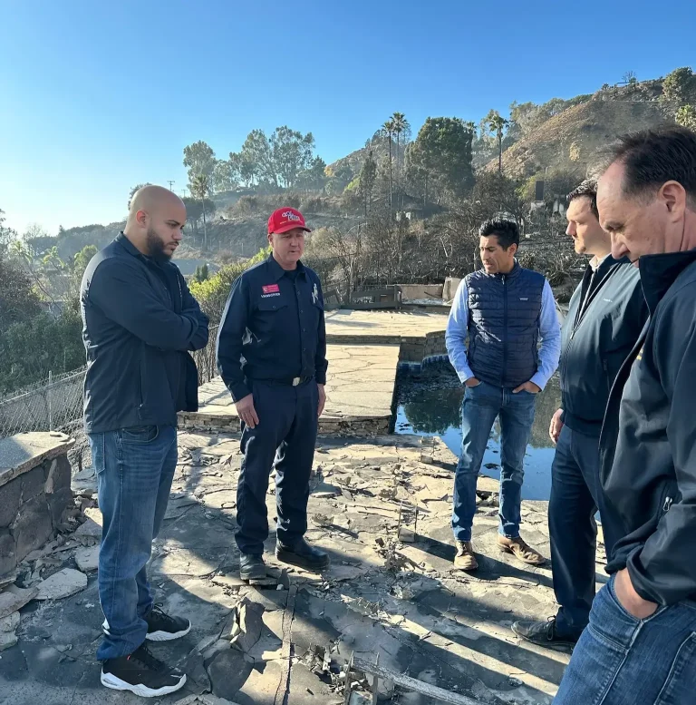 Isaac Bryan, arms folded, listening to a firefighter in a red cap alongside three other men on the fire-scorched patio of a destroyed hillside home, burned slopes rising behind them.