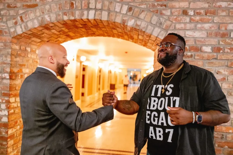 Isaac Bryan in a grey suit clasping hands and laughing with a man in an ‘It’s About Legacy’ T-shirt beneath a brick archway in a public building corridor.
