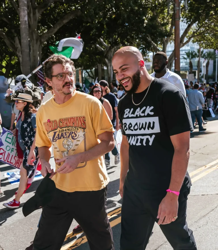 Isaac Bryan laughing as he marches down a city street in a ‘Black Brown Unity’ T-shirt, walking beside another marcher among a crowd carrying flags and signs.