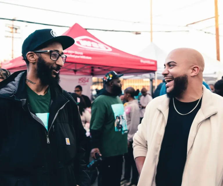 Isaac Bryan in a cream jacket laughing with a man in a cap and glasses at an outdoor community event, vendor canopies and a crowd behind them at dusk.