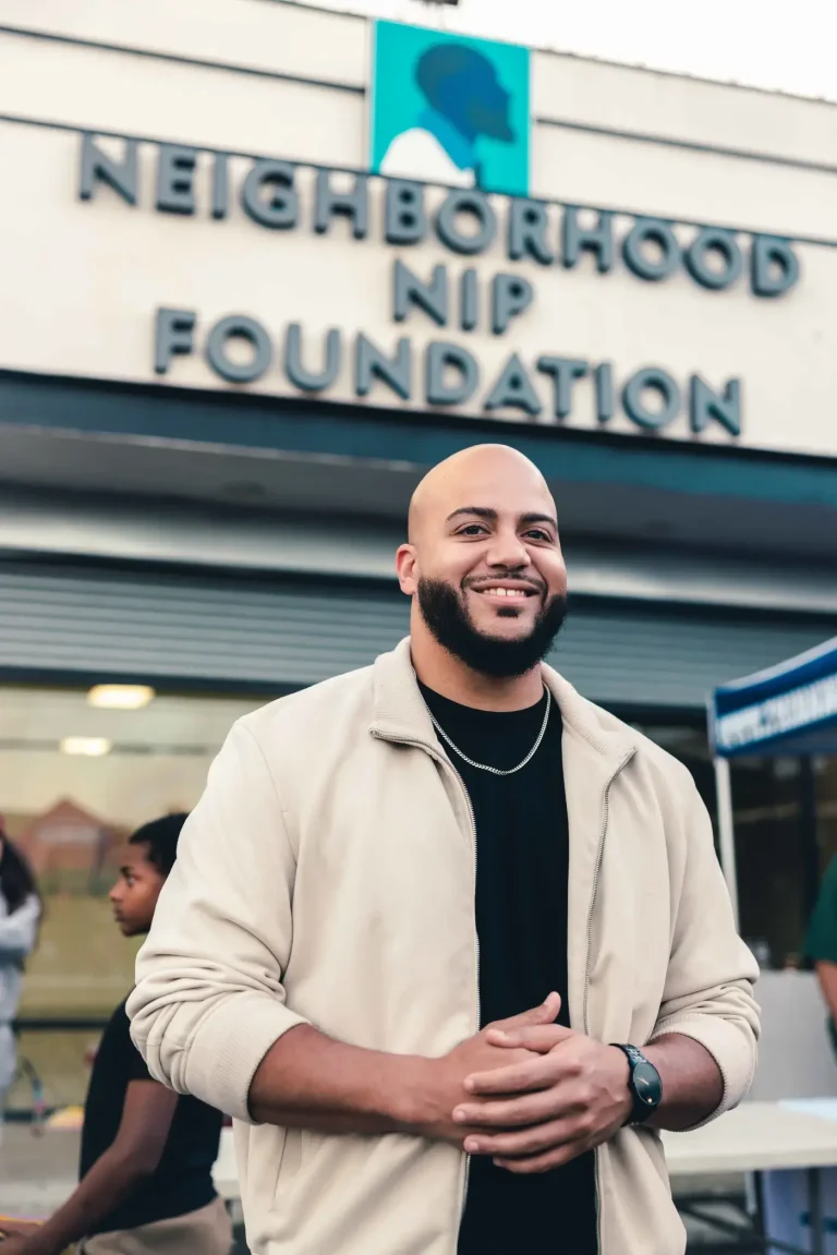 Isaac Bryan smiling with his hands clasped outside the Neighborhood Nip Foundation, the organisation’s raised lettering and logo on the wall above him.