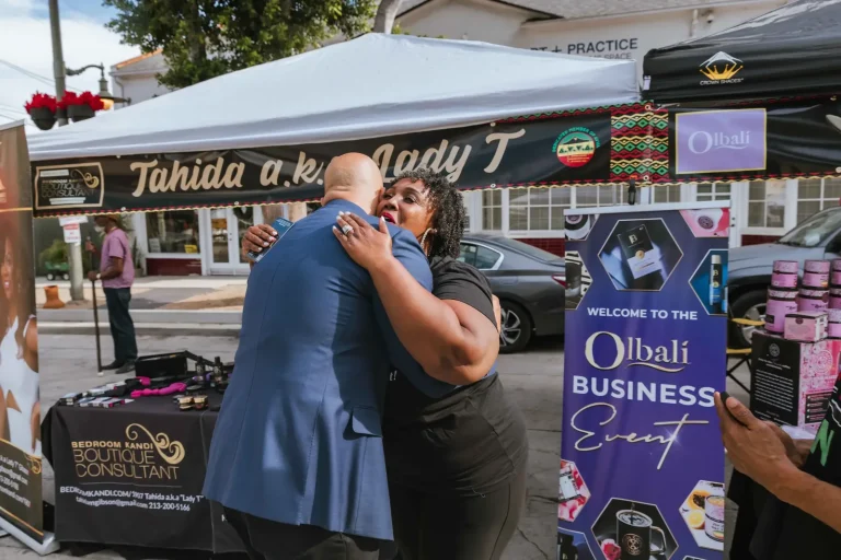 Isaac Bryan, seen from behind in a blue suit, embracing a small-business owner at her vendor booth during an outdoor business showcase, product displays and a welcome banner beside them.