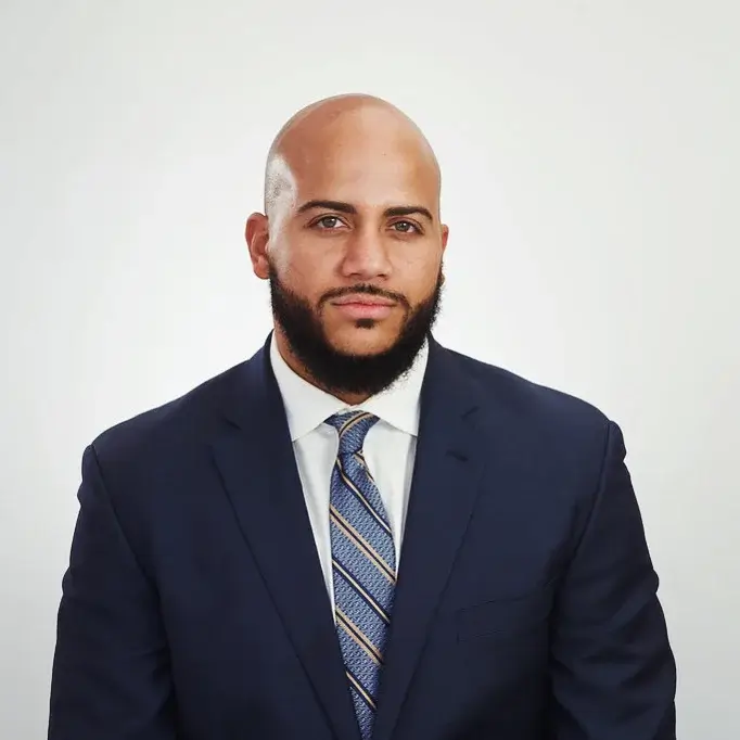 Studio headshot of Isaac Bryan in a navy suit and blue striped tie against a plain light background.