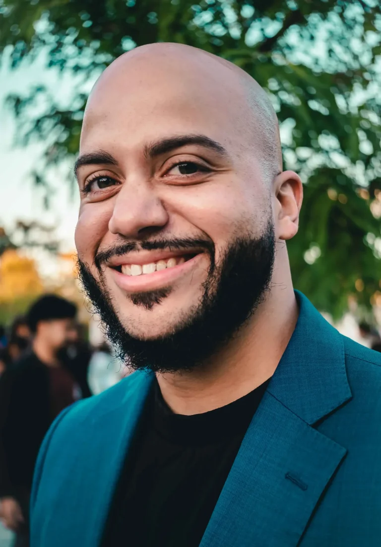 Close-up portrait of Isaac Bryan smiling broadly in a teal blazer at an outdoor evening event, leaves and softly blurred people behind him.