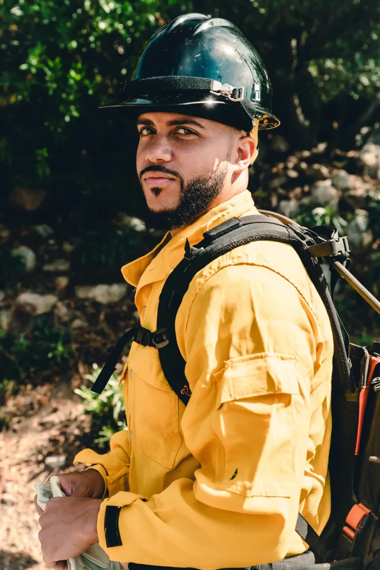 Isaac Bryan in yellow wildland firefighting gear and a hard hat, a pack on his back, looking back over his shoulder on a brush-covered hillside.