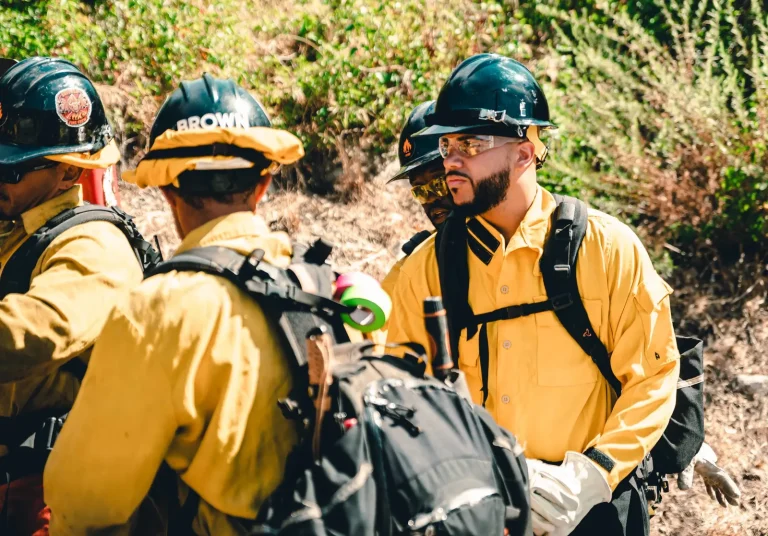 Isaac Bryan in yellow wildland firefighting gear and safety glasses talking with members of a hand crew on a dry hillside, their hard hats and packs in the foreground.