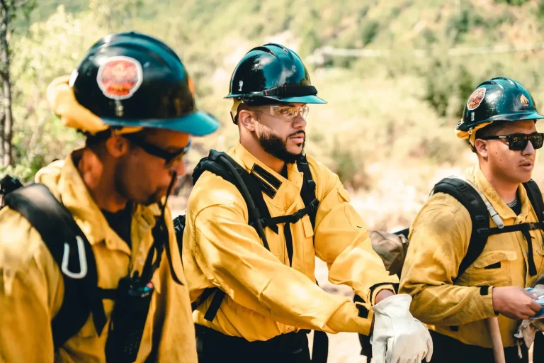 Isaac Bryan standing between two other wildland firefighters in yellow brush gear and hard hats, pulling on a work glove on a sunlit hillside.