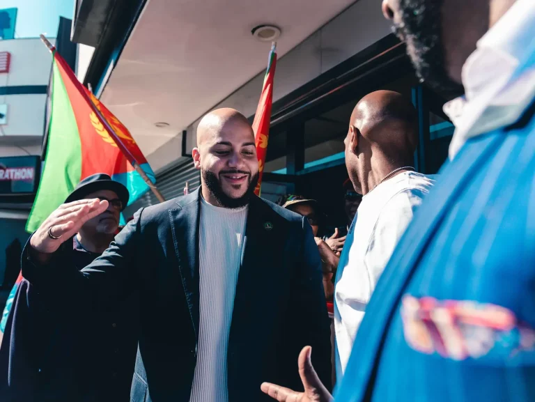 Isaac Bryan smiling as he greets people outside a storefront at a street gathering, Eritrean flags flying behind him.
