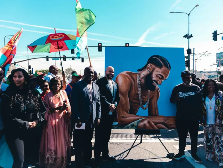 Community members and officials, Isaac Bryan among them in a dark blazer, gathered beside a large painted portrait displayed on an easel at a street intersection, Eritrean flags and a matching parasol raised behind them.