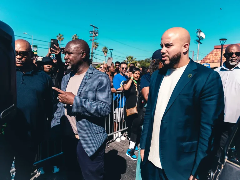 Isaac Bryan in a navy suit walking past metal barricades at a crowded outdoor event, onlookers filming on their phones under a clear blue sky.