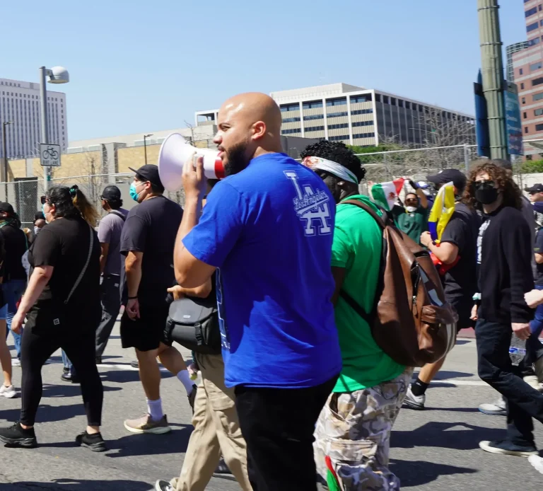Isaac Bryan marching down a downtown Los Angeles street in a blue Dodgers T-shirt, speaking into a megaphone alongside other marchers carrying flags.