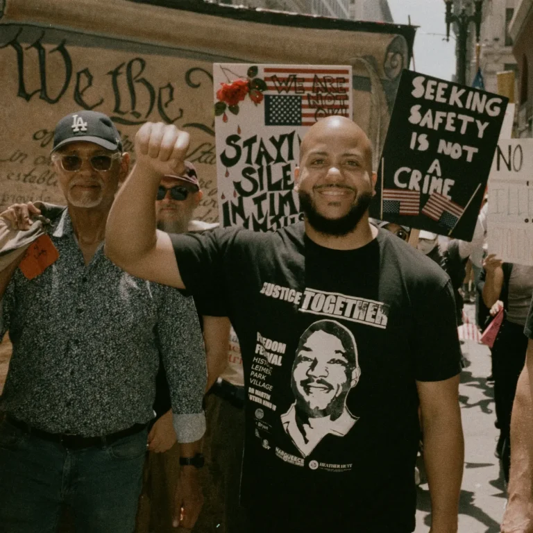 Isaac Bryan smiling with his fist raised at a march, wearing a T-shirt printed with a portrait of Dr Martin Luther King Jr, beside an older man in an LA cap and in front of a large ‘We the People’ Constitution banner and a placard reading ‘Seeking safety is not a crime’.